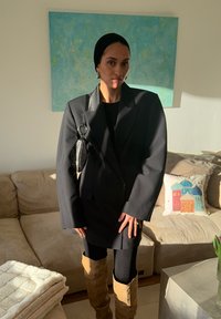 Femme portant un blazer noir surdimensionné, un foulard noir, des leggings noirs, des bottes hautes couleur camel, et un petit sac à main noir, debout dans un salon baigné de lumière.