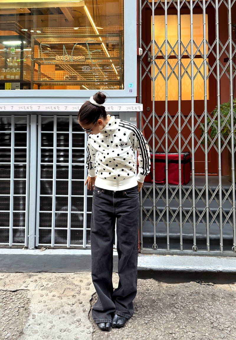 Femme en veste à pois et pantalon large noir, debout et regardant vers le bas devant un magasin fermé avec des grilles en métal et une fenêtre.