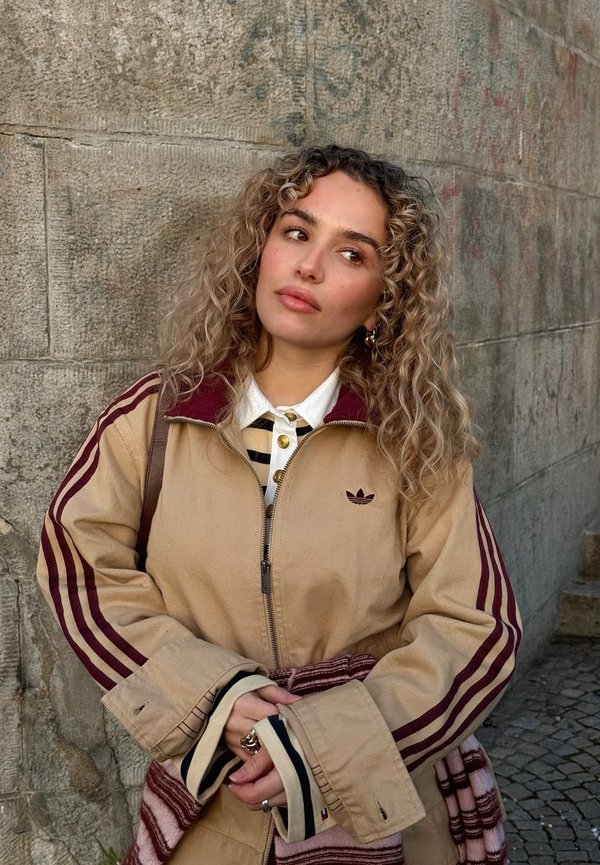 Giovane donna con capelli ricci che indossa una giacca beige con strisce color bordeaux, in piedi contro un muro di pietra strutturato, che guarda di lato, con le mani giunte.