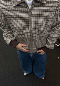 Veste bomber à motif pied-de-poule marron et beige avec une fermeture éclair à l'avant, des poignets côtelés et des poches, associée à un jean bleu à jambes larges et des chaussures blanches.