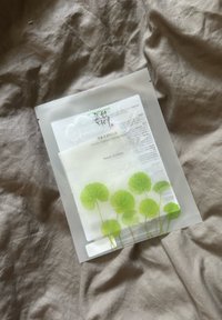 Centella Asiatica raminanti kaukė, pateikta skaidrioje pakuotėje su žaliųjų lapų dizainu priekyje, padėta ant tekstūruoto smėlio spalvos paviršiaus.