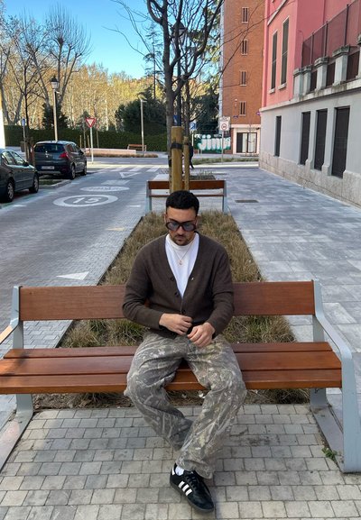 Una persona sentada que lleva un cárdigan marrón sobre una camisa blanca, pantalones de camuflaje y zapatos negros en un banco de madera en un entorno de parque.