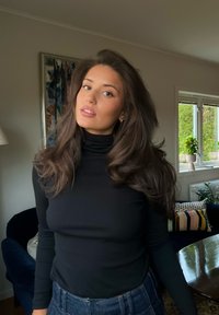 Kobieta z długimi brązowymi włosami, ubrana w czarny golf i niebieskie dżinsy, stoi w salonie z sofą, roślinami i oknem.