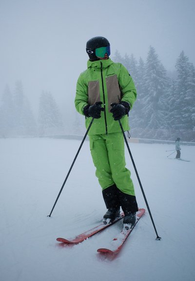 Traje de esquí verde brillante con un panel en el pecho marrón, guantes negros y botas de esquí negras. Los esquís son naranjas. Nieve cayendo en un entorno neblinoso.