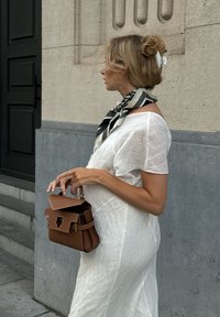 Brown leather handbag featuring a structured design, gold hardware, and a clasp. It is styled with a loose-fitting white dress and a black-and-white patterned scarf.
