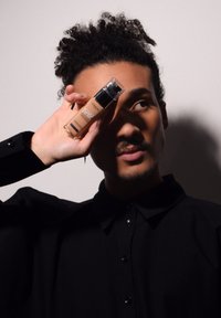 Flacon de fond de teint avec un capuchon pompe, couleur beige, tenu contre une chemise sombre. Texte sur le flacon : "L'Oréal Paris Infallible."