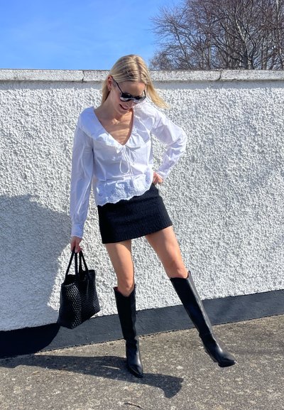 Blusa blanca con ribete de encaje y cordones, combinada con una minifalda de tweed negra, botas negras hasta la rodilla y un bolso negro texturizado.