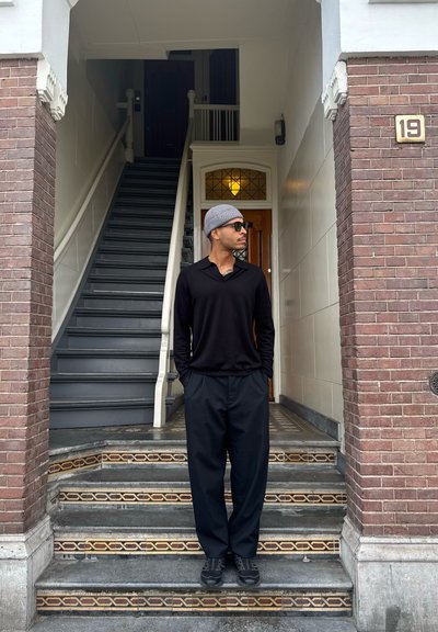 Camisa negra de manga larga, pantalones holgados negros, gorro gris y gafas de sol oscuras; de pie sobre unos escalones de piedra con patrón cerca de una pared de ladrillo.