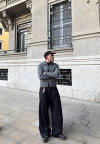 Cardigan grigio sopra una camicia con colletto, pantaloni ampi a gessato nero e berretto nero. Portachiavi visibile sui pantaloni. Sfondo urbano.