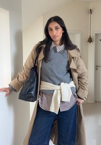 Trench-coat beige par-dessus un pull gris, chemise rayée à col, jean bleu foncé et sac noir. Pull en maille noué autour de la taille.