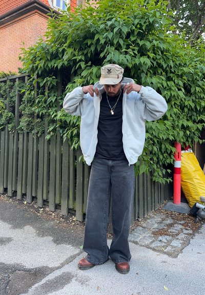 Sudadera gris, camiseta negra, jeans oscuros de pierna ancha y botas marrones. Gorro con patrón de camuflaje. Fondo de follaje verde.