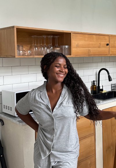 Mujer con pijama gris sonriendo con los ojos cerrados, de pie en una cocina moderna con gabinetes de madera y salpicadero de azulejos blancos.