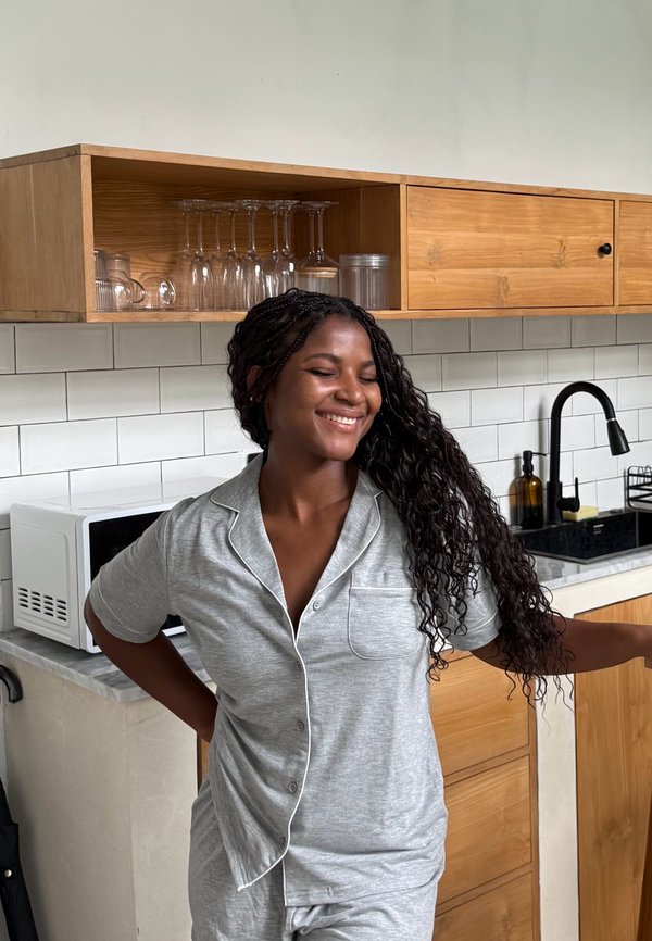 Femme en pyjama gris souriant les yeux fermés, debout dans une cuisine moderne avec des armoires en bois et une crédence carrelée blanche.