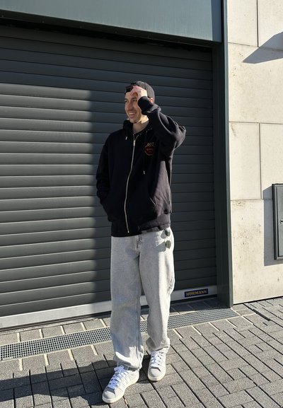 Sudadera negra con cremallera y logo de corazón bordado, pantalones de chándal grises holgados y zapatillas blancas. De pie contra una pared gris texturizada.