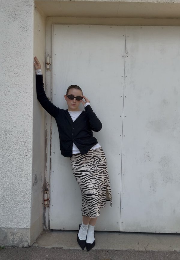 Une femme portant des lunettes de soleil pose devant une porte métallique. Elle porte un cardigan noir, une jupe à imprimé zèbre, des chaussettes blanches et des escarpins pointus.