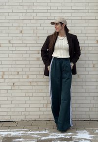 Femme portant une casquette beige, un pull crème, un manteau marron foncé et un pantalon large vert foncé à rayures blanches, debout contre un mur en briques claires.
