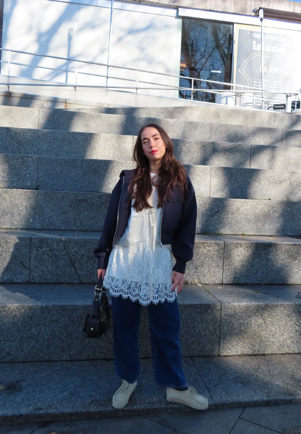 Νεαρή γυναίκα με μακριά μαλλιά, ντυμένη με λευκό δαντελωτό φόρεμα και ναυτικό σακάκι, στέκεται σε πέτρινα σκαλοπάτια κρατώντας μια μαύρη τσάντα χειρός στο φως της ημέρας.