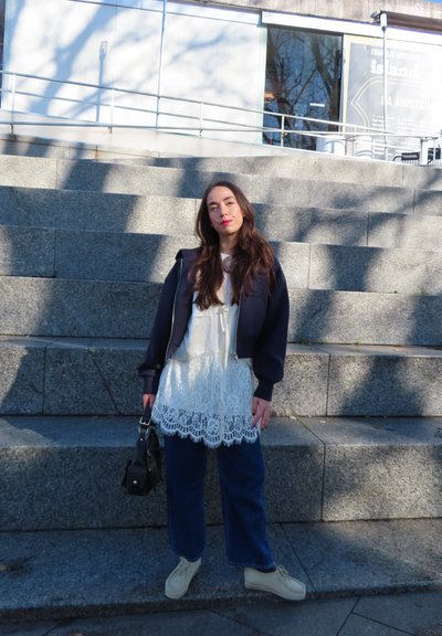 Joven con el cabello largo, vestida con un vestido blanco de encaje y una chaqueta azul marino, de pie sobre unos escalones de piedra sosteniendo un bolso negro a la luz del día.