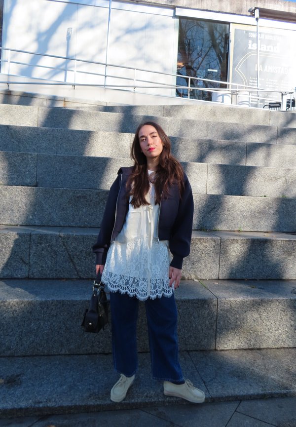 Jonge vrouw met lang haar, gekleed in een witte kanten jurk en een marineblauwe jas, staat op stenen trappen en houdt overdag een zwarte handtas vast.