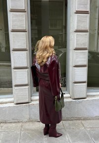 Γυναίκα με μακριά ξανθά μαλλιά που φοράει ένα μπορντό δερμάτινο παλτό και μπότες, κρατώντας μια πράσινη τσάντα, στέκεται σε πεζοδρόμιο από πέτρα κοντά σε ένα παράθυρο.