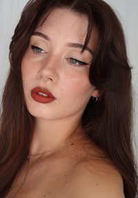Närbild av en person med långt, vågigt mörkt hår, en gyllene septumring, kraftig eyeliner och varm röd läppstift, med svaga fräknar på kinderna.