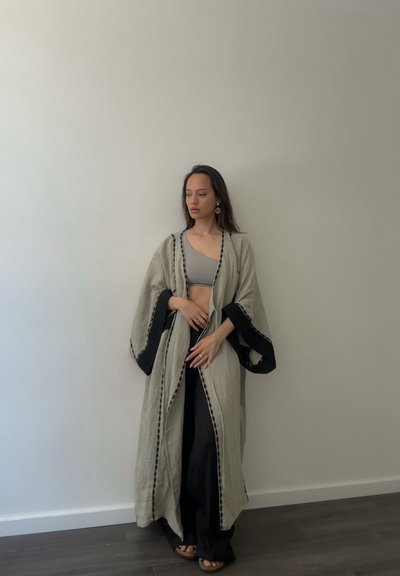 Kimono largo de lino beige con ribete negro y detalles bordados, usado sobre una camiseta gris y pantalones negros; modelo de pie contra una pared lisa.