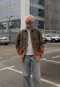 Jeune homme aux cheveux blonds clairs portant une chemise à carreaux brun-orange par-dessus un t-shirt blanc, debout dans un parking urbain.