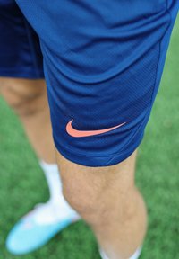 Person som bär blå Nike sportshorts med en rosa logotyp, står på grön gräs, med ett suddigt ben och en blå fotbollssko synlig.