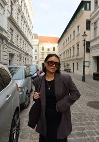 Per didelės apimties pilka švarkas, juoda palaidinė ir sijonas. Rudas raštuotas rankinukas ir dideli akiniai nuo saulės. Akmenimis grįstas takas su šalia parkavais automobiliais.