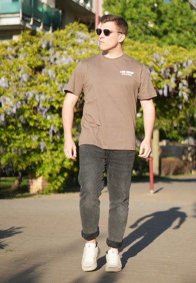 Hombre con gafas de sol, camiseta marrón, vaqueros oscuros y zapatillas blancas caminando al aire libre en un día soleado con follaje verde de fondo.