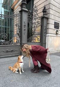 Un Shiba Inu avec un collier rouge est assis sur un trottoir gris à côté d'une femme portant un long manteau bordeaux, des collants à motifs et des bottes marron, se penchant en avant.