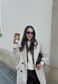 Femme en manteau beige et lunettes de soleil tenant un gobelet à couvercle rouge avec un motif de Noël et un bonhomme en pain d'épice, debout dans une rue pavée.