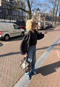 Črna majica s napihnjenimi rokavi, hlače z visokim pasom v blede barvi, mrežasti superge, drži vzorčasto torbico z risanim junakom, na obdani ulici iz opeke.