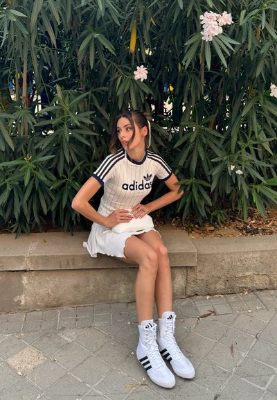 Camiseta deportiva blanca de Adidas con rayas negras, combinada con una falda blanca y zapatillas altas blancas de Adidas con acentos negros.