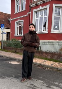 Ruda bombonešio striukė su tamsia skarele, juodos platėjamos kelnės ir rudi batai su storu padu, stovintis gatvėje prie raudono pastato.