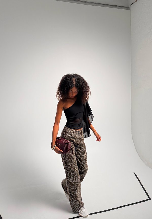 Mujer con pelo rizado vestida con un top negro de un solo hombro y pantalones con estampado de leopardo, sosteniendo un bolso burdeos, de pie sobre un suelo blanco de estudio.