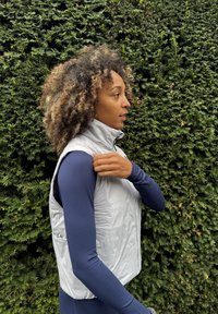 Viegls sudraba bezpiedurkņu veste ar augstu apkakli; ar rāvējslēdzēja aizdari un sānu kabatām; valkāta virs gar�ām piedurknēm tumši zils tops.