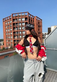 Svart keps med "OUTLAWS"-logotyp, röd och vit oversized jacka, svart bralette, röd rutiga slips, grå trasiga sweatpants, texturerad urban bakgrund.