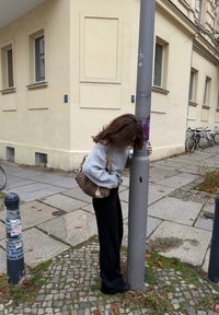 Pull gris, pantalon noir et sac marron à motifs ; personne appuyée contre un poteau gris dans une rue de la ville pavée avec des vélos.