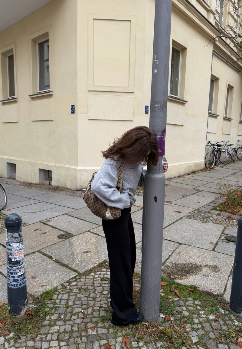 Pull gris, pantalon noir et sac marron à motifs ; personne appuyée contre un poteau gris dans une rue de la ville pavée avec des vélos.