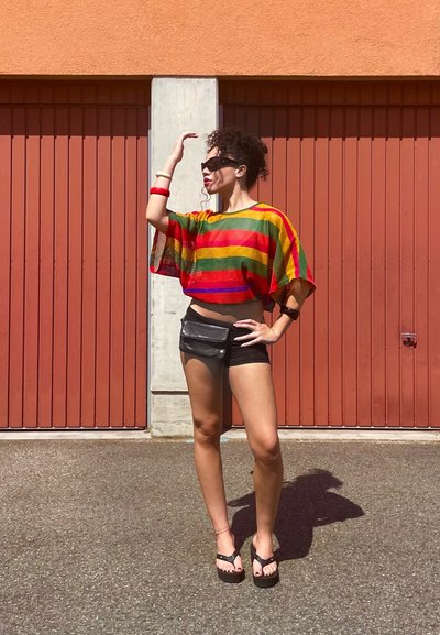 Top corto de rayas multicolores con tonos rojos, verdes y amarillos, combinado con pantalones cortos negros y una bolsa de cintura oscura, contra un fondo rojo.