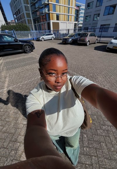 Una persona con el cabello corto y peinado hacia atrás lleva gafas redondas y una camisa verde claro. Está de pie en un área pavimentada con coches aparcados y edificios.