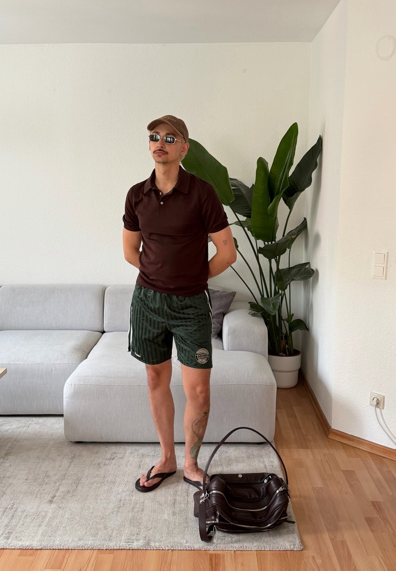 Homme portant des lunettes de soleil, une casquette, une chemise sombre et un short, debout pieds nus près d'un canapé et d'une grande plante d'intérieur dans un salon avec un sol en bois.