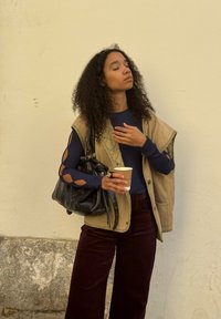 Fiatal nő göndör hajjal áll a falnak támaszkodva, csukott szemmel, kávéscsészét és fekete kézitáskával a kezében, réteges ruházatban.