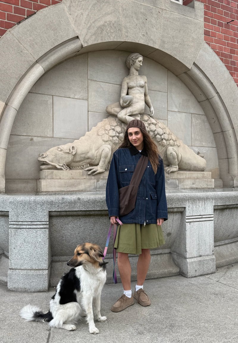 Junge Frau in Jeansjacke und grünem Rock hält eine Hundeleine und steht vor einer Steinskulptur einer sitzenden Figur auf einem Reptil.
