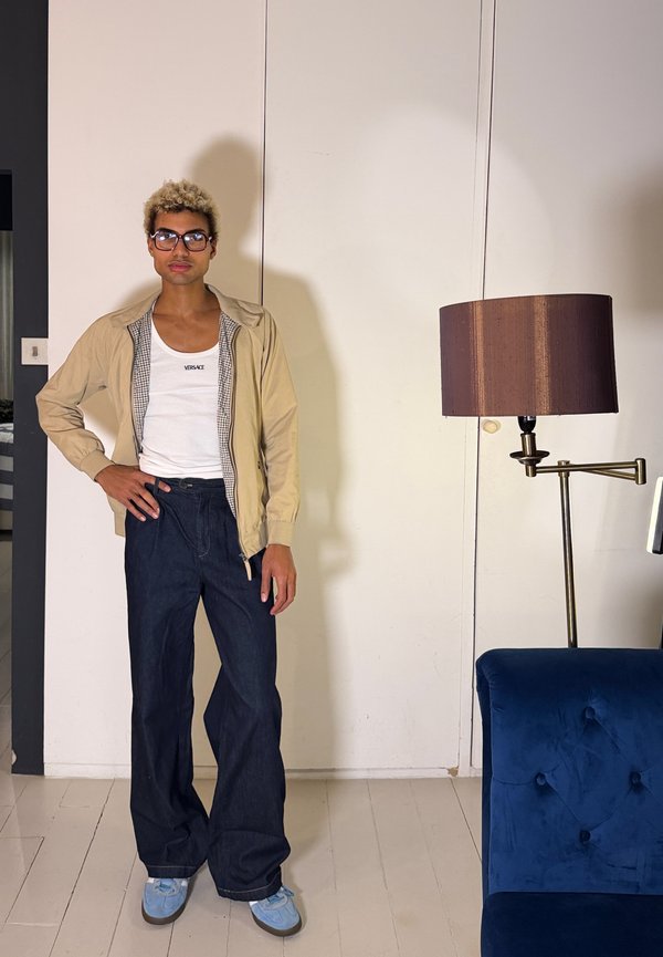 Rövid göndör szőke hajú, szemüveges személy bézs dzsekiben, fehér Versace trikóban, széles sötét farmernadrágban és kék tornacipőben, bent áll egy lámpa és kanapé közelében.