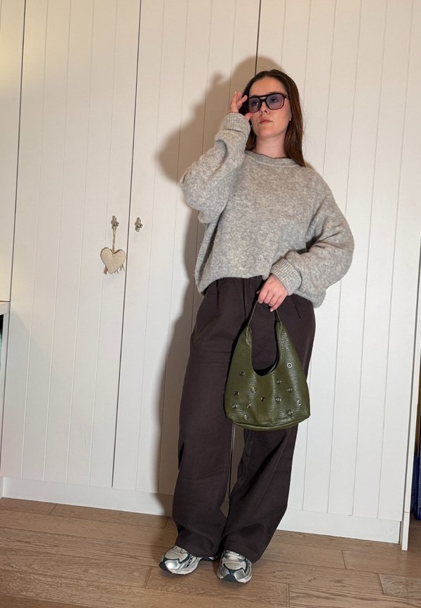 Femme portant un pull gris oversize, un pantalon ample foncé, des baskets et des lunettes de soleil, tenant un sac à main vert clouté à l'intérieur.