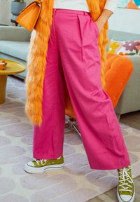 Άτομο που φοράει φωτεινό ροζ παντελόνι με φαρδιά πόδια, πράσινα sneakers ψηλά και πορτοκαλί γούνινο παλτό, στέκεται σε ένα πολύχρωμο σαλόνι.