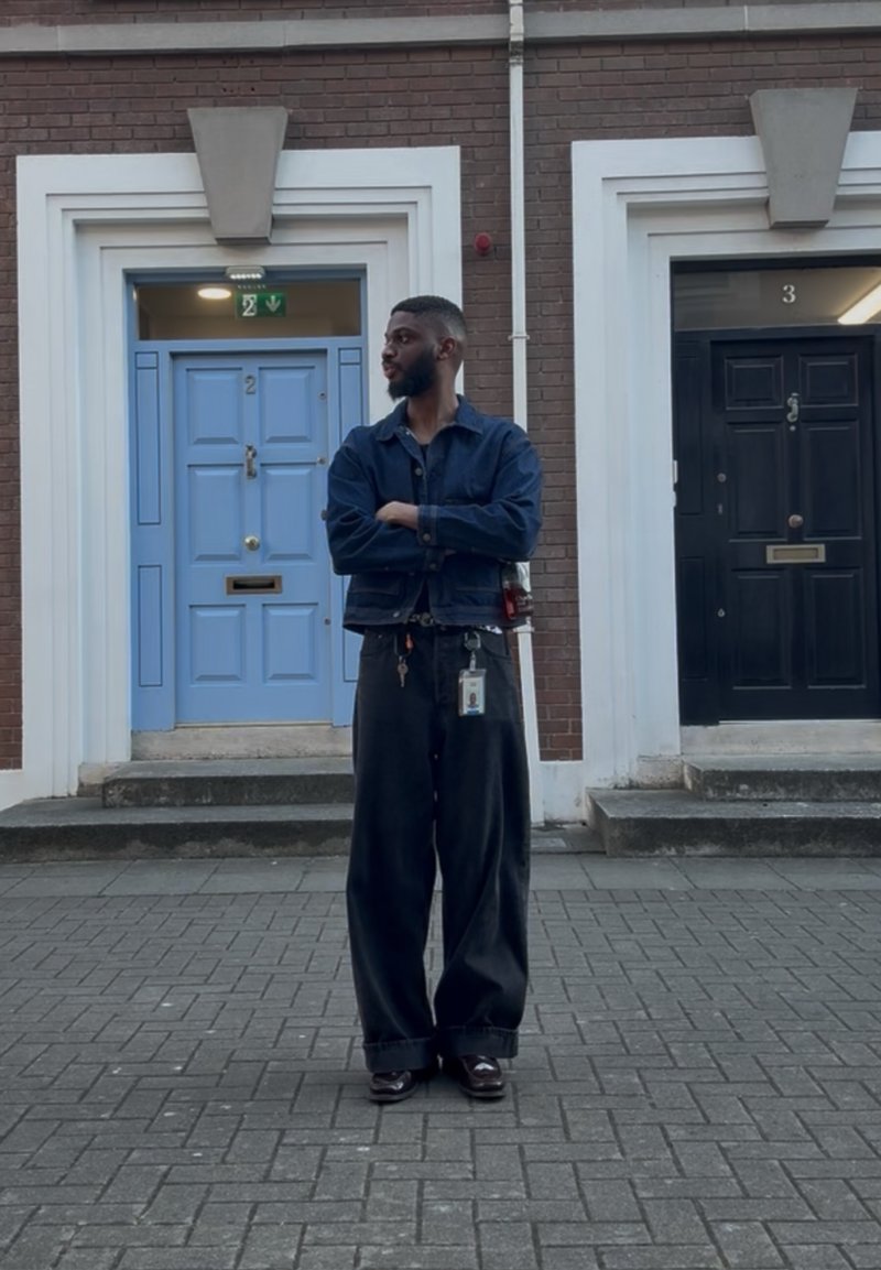 Homme avec barbe portant un pantalon large et sombre et une veste, debout les bras croisés dans la rue entre deux portes numérotées 2 et 3.