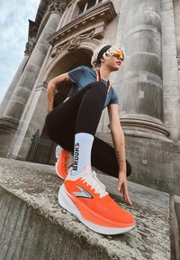 Zapatillas de running de un vibrante color naranja con suelas blancas y la marca "Brooks". La parte superior tiene un acabado texturizado; los cordones son de color naranja neón.
